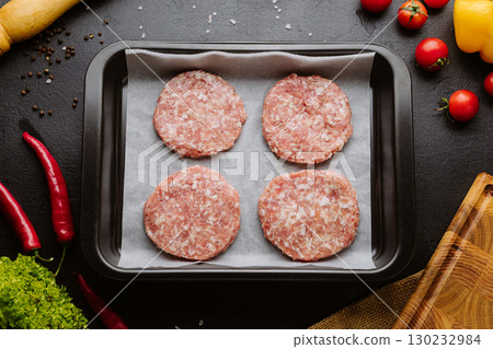 Raw burger patties on baking tray with fresh vegetables and spices on dark background 130232984
