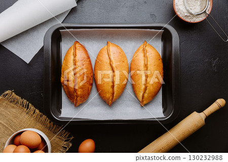 Golden homemade pies in baking tray, rustic kitchen scene with rolling pin and eggs 130232988