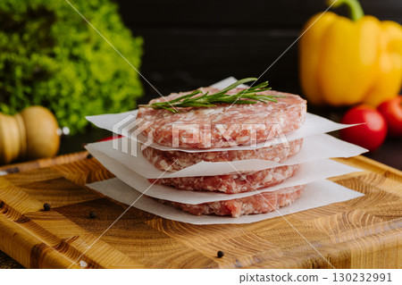 Raw burger patties stacked with parchment and rosemary on a wooden board, ready to cook 130232991