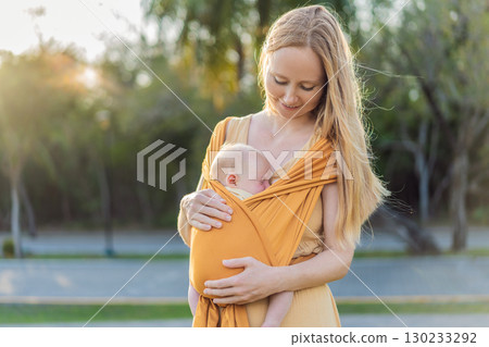 Mother holding her little baby in a yellow sling in the park. Warm and loving family moment. Babywearing, parenting, and mother-child bonding concept 130233292