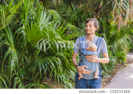 Young father walks in nature with his baby son nestled close in a parent-facing ergonomic baby carrier. Bonding, warmth, and modern parenting concept in a peaceful outdoor setting 130233369