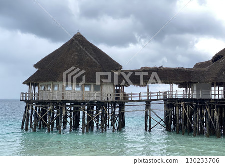A picturesque wooden pier and restaurant with thatched roofs extend over the turquoise waters of the Indian Ocean, seen from a pristine white-sand beach 130233706