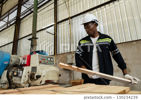 African American male worker in protective gear operates a woodworking machine and inspects lumber planks in a factory, representing precision, manufacturing, and safety. African American male worker in protective gear operates a woodworking machine and inspects lumber planks in a factory, representing precision, manufacturing, and safety. 130234239