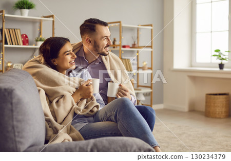 Loving affectionate couple sitting in modern living room wrapped with warm blanket, happy romantic  130234379