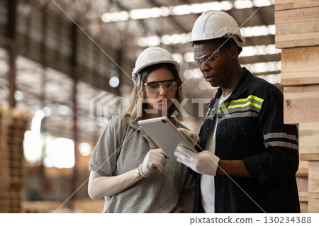 Two Multi-ethnic industrial workers wearing safety helmets and gloves use digital tablet for data analysis in warehouse, showcasing teamwork, technology wooden manufacturing processes. Two Multi-ethnic industrial workers wearing safety helmets and gloves use digital tablet for data analysis in warehouse, showcasing teamwork, technology wooden manufacturing processes. 130234388