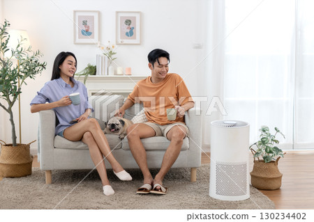 young asian couple drinking morning coffee sitting on sofa relaxing time living room with small pug dog rests beside them with air purifier for clean air in house for good health. 130234402