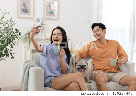 Happy young asian couple drinking morning coffee sitting on sofa relaxing time living room smiling use smartphone take selfies photo with small pug dog rests beside. Happy young asian couple drinking morning coffee sitting on sofa relaxing time living room smiling use smartphone take selfies photo with small pug dog rests beside. 130234445
