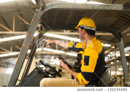 Industrial worker wearing safety helmet and uniform pointing and communicating instructions while operating forklift in warehouse. Team coordination and logistics planning in action. 130234499