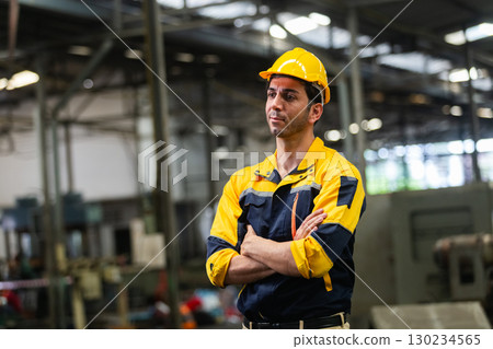 Industrial engineer with crossed arms standing in factory, wearing safety helmet and uniform, expressing confidence, responsibility, focus, and leadership in a manufacturing environment. Industrial engineer with crossed arms standing in factory, wearing safety helmet and uniform, expressing confidence, responsibility, focus, and leadership in a manufacturing environment. 130234565