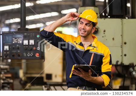 Smiling industrial engineer wearing safety helmet and holding clipboard while standing in factory, representing workplace inspection, manufacturing operations and engineering management. Smiling industrial engineer wearing safety helmet and holding clipboard while standing in factory, representing workplace inspection, manufacturing operations and engineering management. 130234574