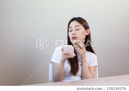 Asian woman examining her face and checking skin problem with compact mirror, showing concern about acne, skincare routine, and facial beauty treatment at home. 130234576