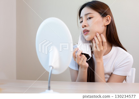 Asian woman removing face mask and checking acne in mirror with concern, representing mask problem, skincare issue, and facial irritation caused by prolonged mask use. 130234577