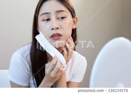 Asian woman removing face mask and checking acne in mirror with concern, representing mask problem, skincare issue, and facial irritation caused by prolonged mask use. 130234578