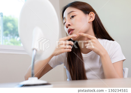 Asian woman checking acne on her face in mirror and squeezing pimple, showing skincare concern, beauty problem, and facial treatment routine for clear healthy skin. 130234581