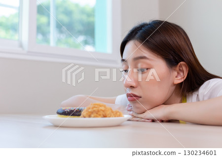 Asian woman staring at fried chicken and a donut with hesitation, representing diet struggle, food temptation, and healthy eating challenge. Ideal for concepts of nutrition decisions and weight Asian woman staring at fried chicken and a donut with hesitation, representing diet struggle, food temptation, and healthy eating challenge. Ideal for concepts of nutrition decisions and weight 130234601