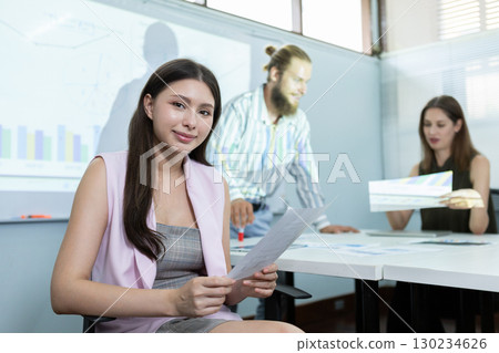 Confident young woman holding business report in meeting room with team discussing data analytics in background, representing leadership, teamwork, and corporate presentation success. 130234626