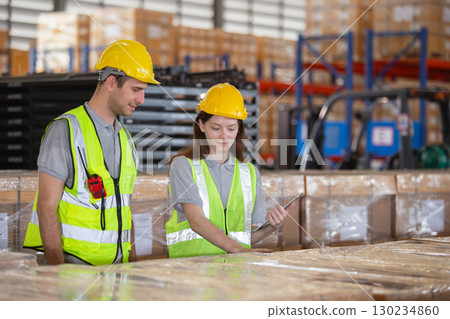 Warehouse workers in safety vests and helmets inspecting inventory and using digital tablets for stock management. Teamwork in modern logistics and industrial operations. 130234860