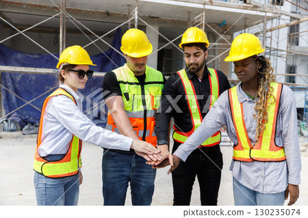 Diverse construction team wearing safety helmets and reflective vests stacking hands together at building site, representing unity, teamwork, collaboration and commitment to project success. 130235074