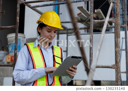 Female construction engineer wearing safety helmet and reflective vest using digital tablet at building site, representing technology, project management, safety and engineering solutions. Female construction engineer wearing safety helmet and reflective vest using digital tablet at building site, representing technology, project management, safety and engineering solutions. 130235148