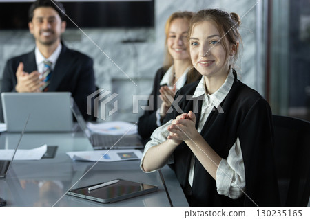 Smiling businesswoman in conference room applauding during a meeting, showing teamwork, support, and positive communication in a modern corporate environment. 130235165