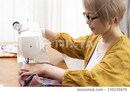 一位老年婦女高興地用縫紉機縫製衣服和配件 一位老年婦女高興地用縫紉機縫製衣服和配件 130239870