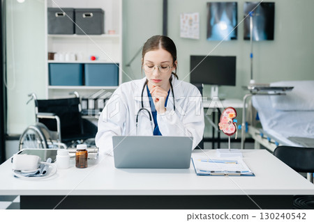 Confident young Caucasian female doctor in white medical uniform sit at desk working on computer. Smiling use laptop write in medical journal 130240542