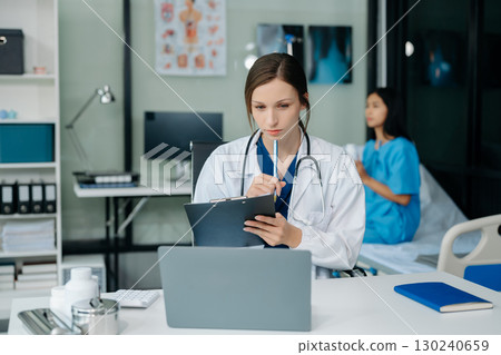 Confident young Caucasian female doctor in white medical uniform sit at desk working on computer. Smiling use laptop write in medical journal 130240659