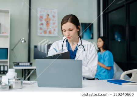 Confident young Caucasian female doctor in white medical uniform sit at desk working on computer. Smiling use laptop write in medical journal 130240660