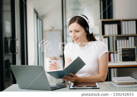 happy young student studying at the college library, sitting at the desk, using a laptop computer, tablet and headphones having a video chat. 130242655