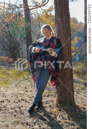 Portrait of smiling caucasian girl plays ukulele in autumnal park. Young woman plays guitar musical instrument outside in nature fall time. Audio music healing Portrait of smiling caucasian girl plays ukulele in autumnal park. Young woman plays guitar musical instrument outside in nature fall time. Audio music healing 130243712