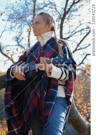 Beautiful blonde girl playing guitar ukulele and singing relaxing enjoying outdoors on sunny autumn day. Hippie woman in boho style training playing musical instrument. Fall leaves 130243728