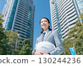 a japanese female engineer wearing a blue suit and holding a white helmet stands in front of tall office buildings 130244236