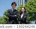 a young japanese businessman and woman wearing black suits are smiling while standing in front of an office building 130244246