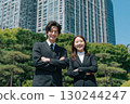 a young japanese businessman and woman wearing black suits are smiling while standing in front of an office building 130244247