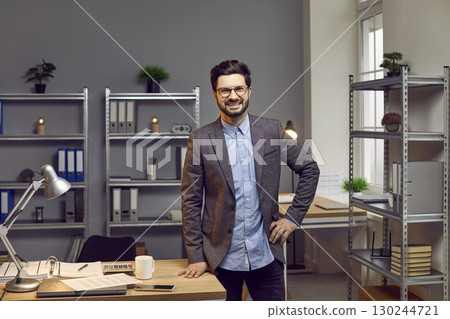 Portrait of smiling, happy, self-confident young businessman with glasses leaning on desk with stationery in spacious creative office. Portrait of smiling, happy, self-confident young businessman with glasses leaning on desk with stationery in spacious creative office. 130244721