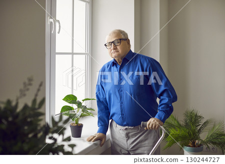 Senior man standing at home in rehabilitation at the window with his crutch and looking at camera. Senior man standing at home in rehabilitation at the window with his crutch and looking at camera. 130244727