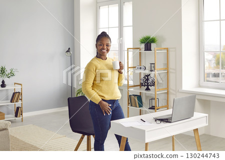Beautiful smiling African American woman drinks from a cup standing in a cozy room. 130244743