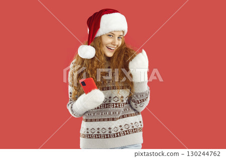 Happy lucky girl in Santa hat is showing yes gesture holding smartphone on red background. 130244762