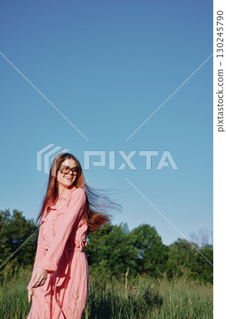 Smiling young woman in a pink dress enjoying a sunny day outdoors with flowing hair and a clear blue sky, evoking joy and freedom in nature 130245790