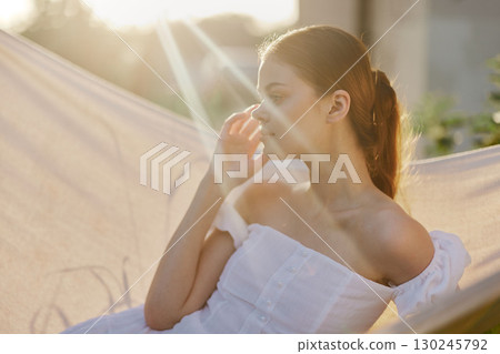 A contemplative young woman in a white off shoulder dress, enjoying a serene moment in a sunlit hammock, surrounded by greenery during golden hour 130245792