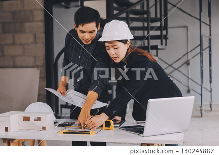 Construction manager and engineer dressed in orange work vests and hard helmets explore construction documentation on the building site near the steel frames 130245887