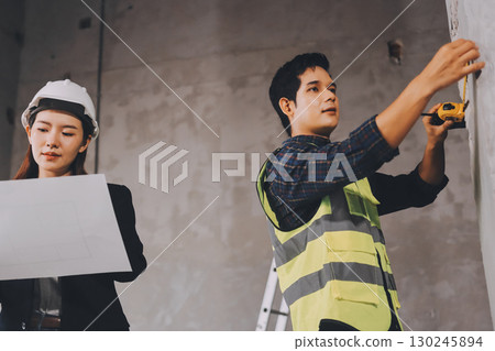 Construction manager and engineer dressed in orange work vests and hard helmets explore construction documentation on the building site near the steel frames 130245894