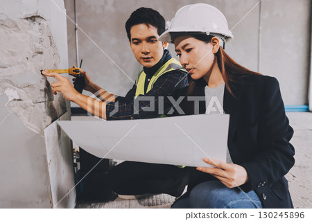 Construction manager and engineer dressed in orange work vests and hard helmets explore construction documentation on the building site near the steel frames 130245896