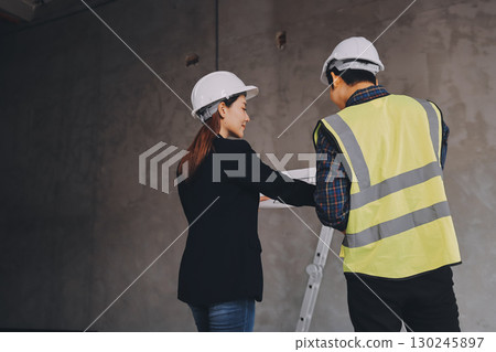 Construction manager and engineer dressed in orange work vests and hard helmets explore construction documentation on the building site near the steel frames 130245897