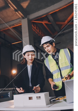 Construction manager and engineer dressed in orange work vests and hard helmets explore construction documentation on the building site near the steel frames 130245901