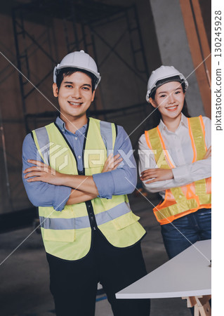 Construction manager and engineer dressed in orange work vests and hard helmets explore construction documentation on the building site near the steel frames Construction manager and engineer dressed in orange work vests and hard helmets explore construction documentation on the building site near the steel frames 130245928