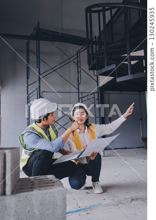 Construction manager and engineer dressed in orange work vests and hard helmets explore construction documentation on the building site near the steel frames Construction manager and engineer dressed in orange work vests and hard helmets explore construction documentation on the building site near the steel frames 130245935