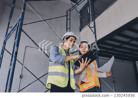 Construction manager and engineer dressed in orange work vests and hard helmets explore construction documentation on the building site near the steel frames Construction manager and engineer dressed in orange work vests and hard helmets explore construction documentation on the building site near the steel frames 130245936