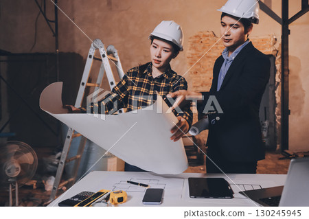 Construction manager and engineer dressed in orange work vests and hard helmets explore construction documentation on the building site near the steel frames Construction manager and engineer dressed in orange work vests and hard helmets explore construction documentation on the building site near the steel frames 130245945