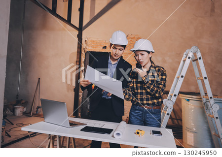 Construction manager and engineer dressed in orange work vests and hard helmets explore construction documentation on the building site near the steel frames 130245950